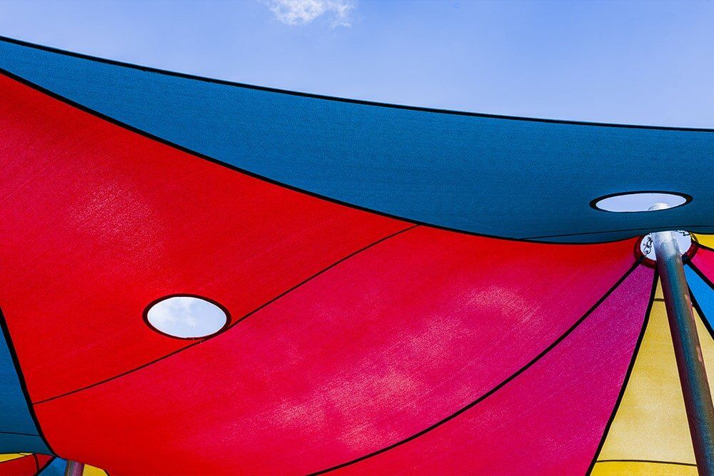 Tensioned red, teal and yellow waterproof shade sails with ventilation eyelets against a blue sky